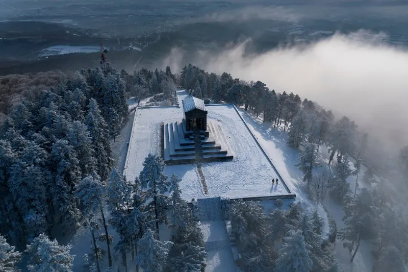 An aerial view of the black granite Monument to the Unknown Hero on the top of Mount Avala near Belgrade, among snow-covered pine trees and morning fog.