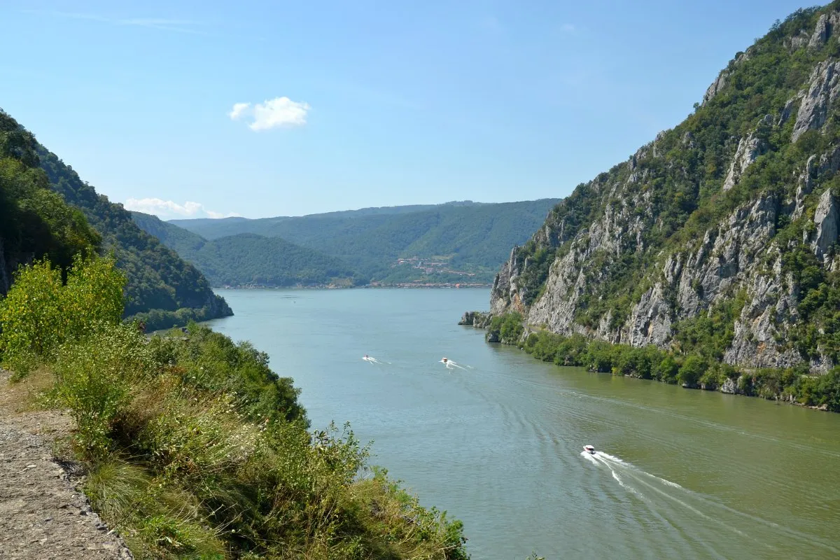 The Danube winds between steep wooded cliffs in the Iron Gates gorge between Serbia and Romania, with small boats on the green water.