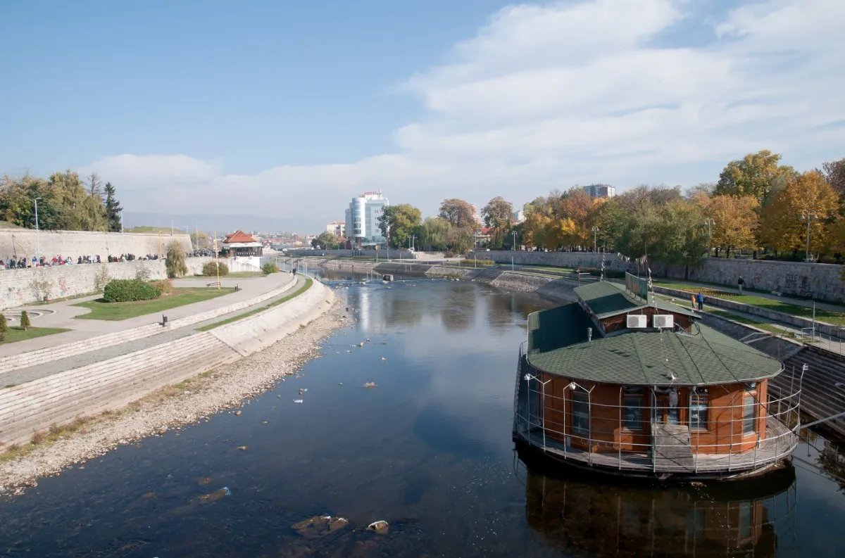 The Nišava river flows between stone embankments through the centre of Niš, with a round floating house on the water and autumn trees along the bank.