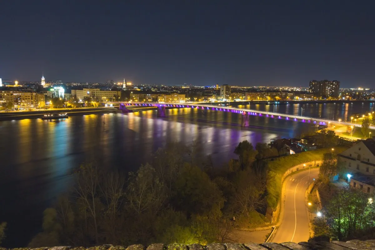 The lights of Novi Sad and the purple-lit Varadin Bridge reflected in the Danube at night, seen from the Petrovaradin Fortress.