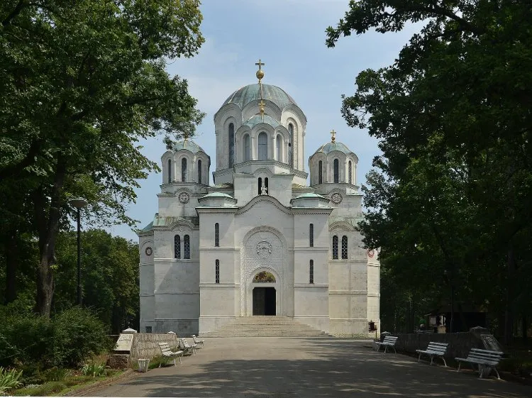 The white marble St. George's Church on Oplenac hill in Topola with its green domes, at the end of a paved path between tall trees.
